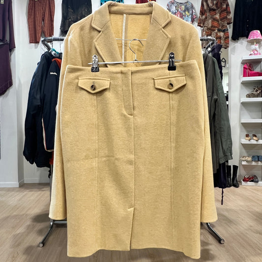 Front view of a beige wool skirt suit set with a knee-length skirt featuring buttoned flap pockets, displayed together with the matching blazer on a hanger inside a vintage clothing store