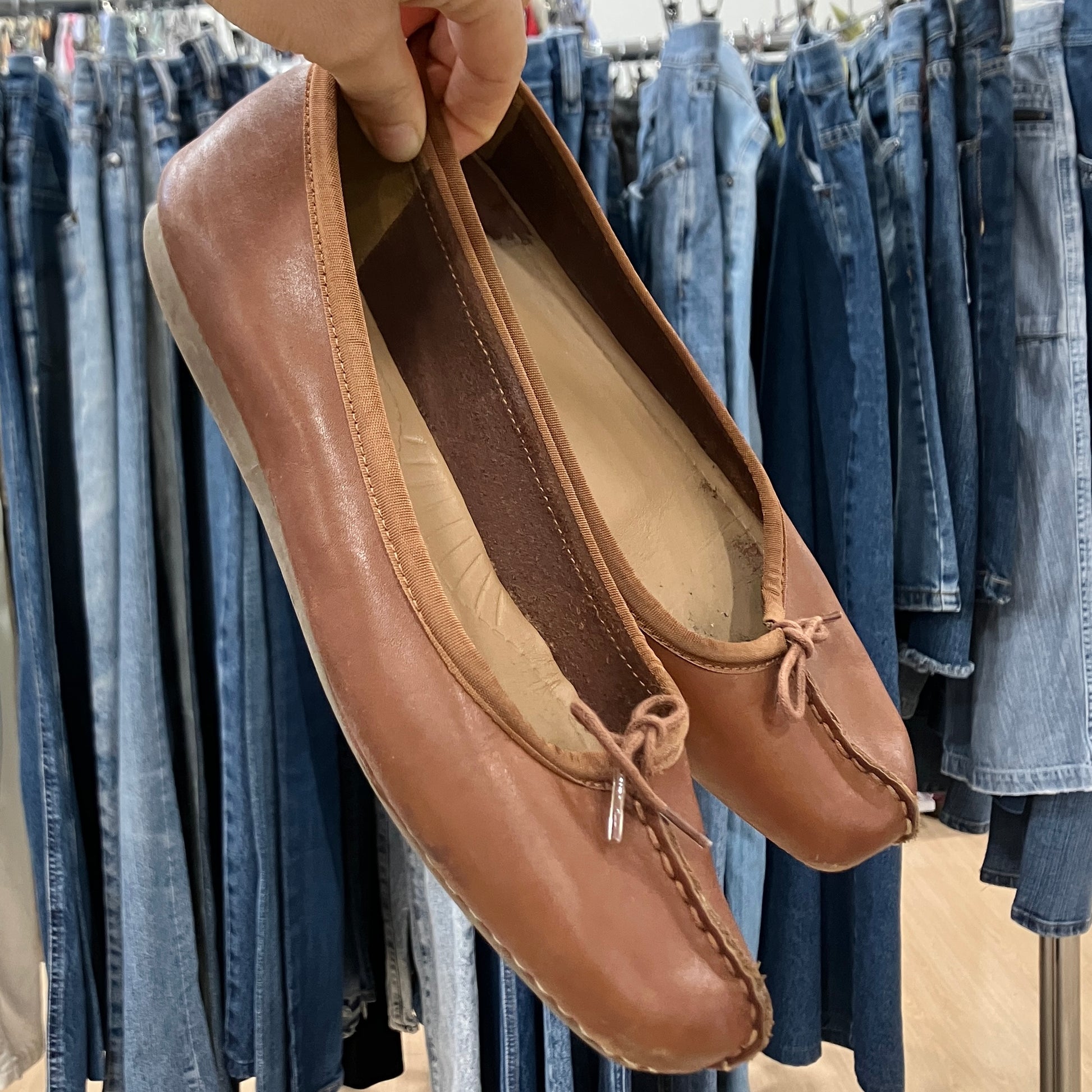 Brown leather shoes held up in front of a clothing rack with jeans